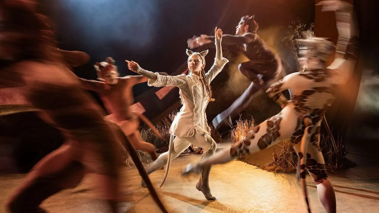 Central dancer in beige tunic with cat ears leaps forward while other performers blur around on a dim, dramatic stage setup.