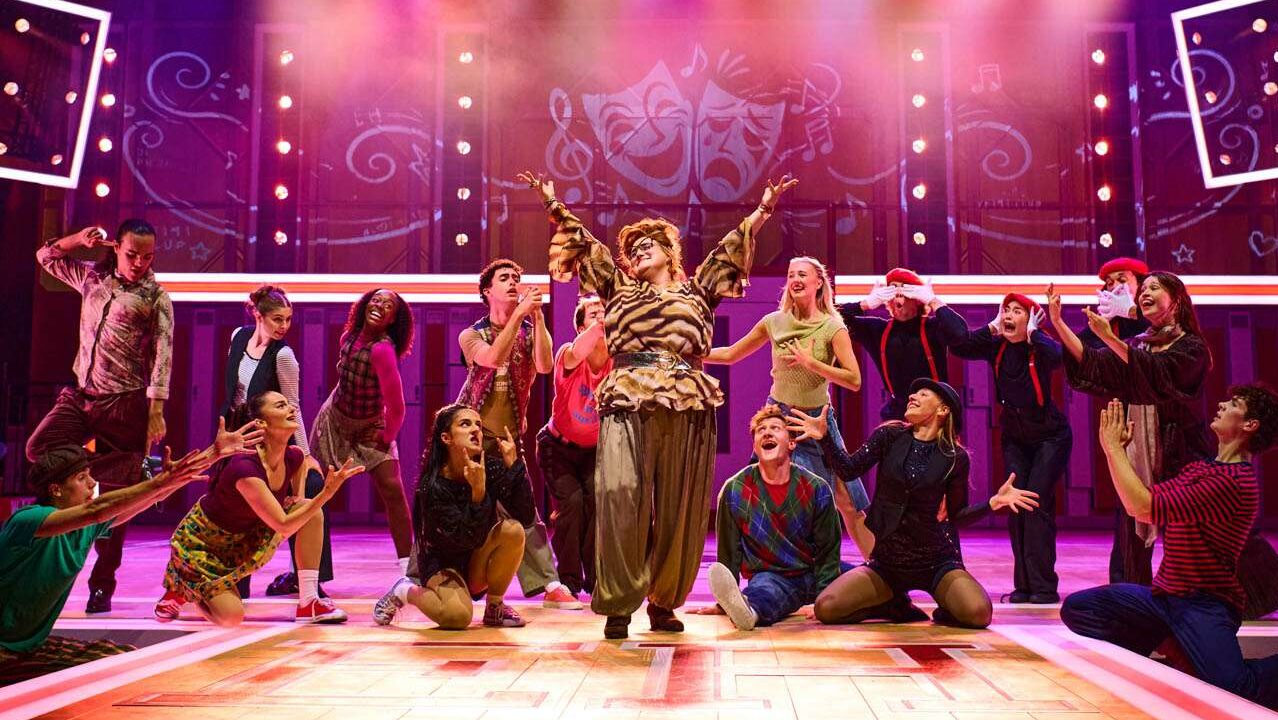 Diverse group of dancers on a brightly lit stage, central performer raises arms as others pose around them.