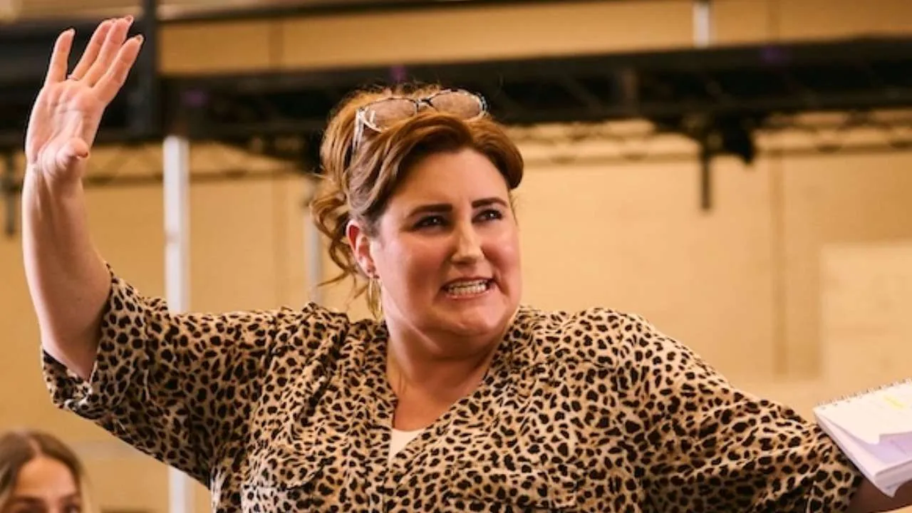Woman in a leopard-print blouse raises her hand while speaking in a meeting or workshop, with notes in hand.