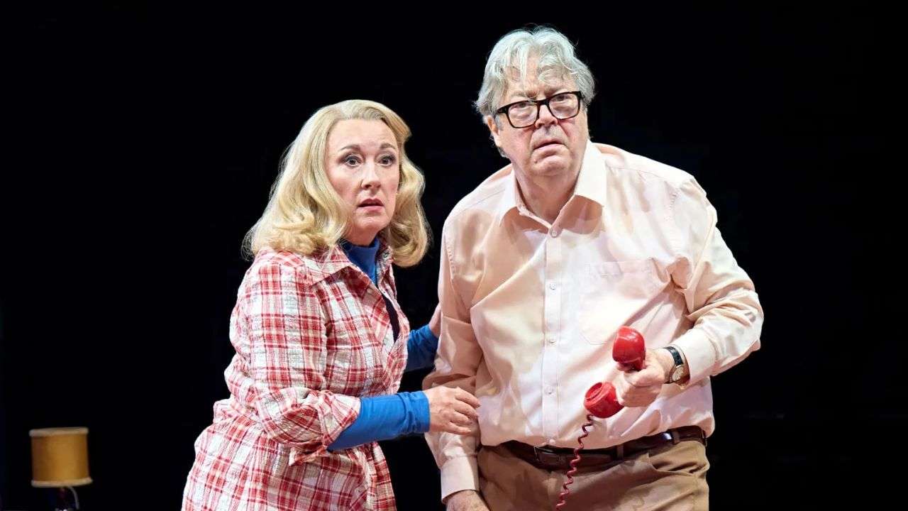Two actors on a dark stage, the man holding a red corded phone and both looking surprised or concerned.