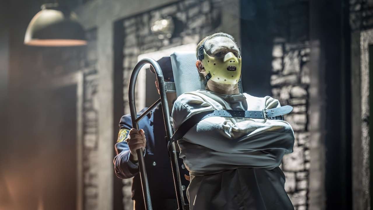 Person in a restraint chair wearing a pale hockey-style mask in a dim, industrial-looking room.