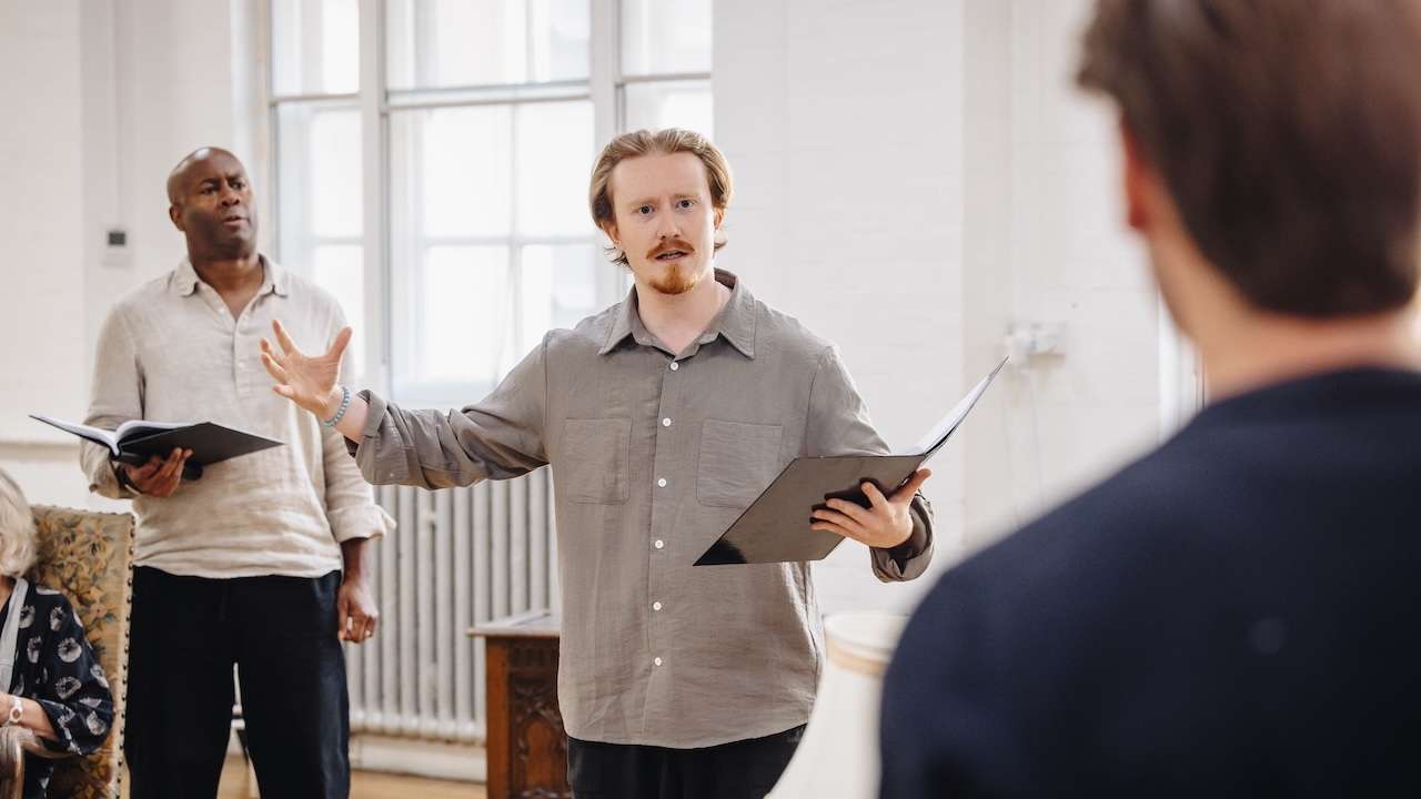 Man in a gray shirt holds a folder and gestures as he speaks to two others in a bright, spacious room.