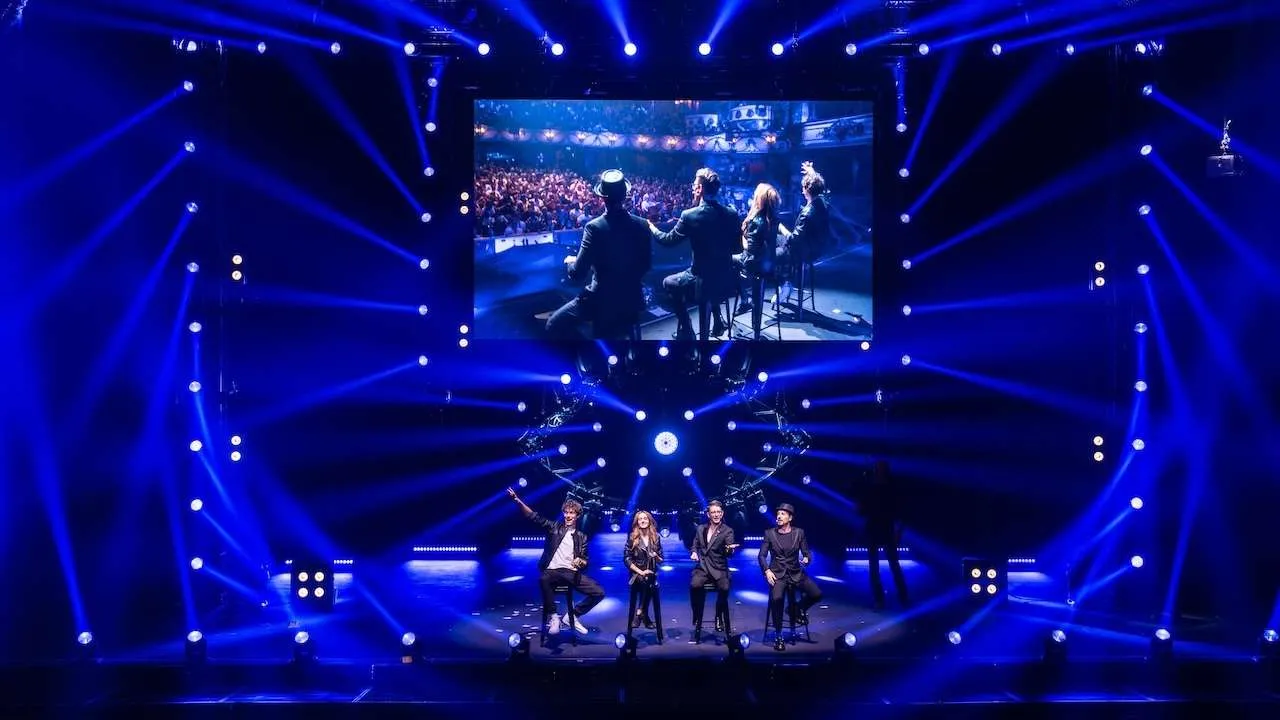 Stage performance with five singers seated center, blue spotlight beams sweeping the venue, and a large screen showing the audience above them.