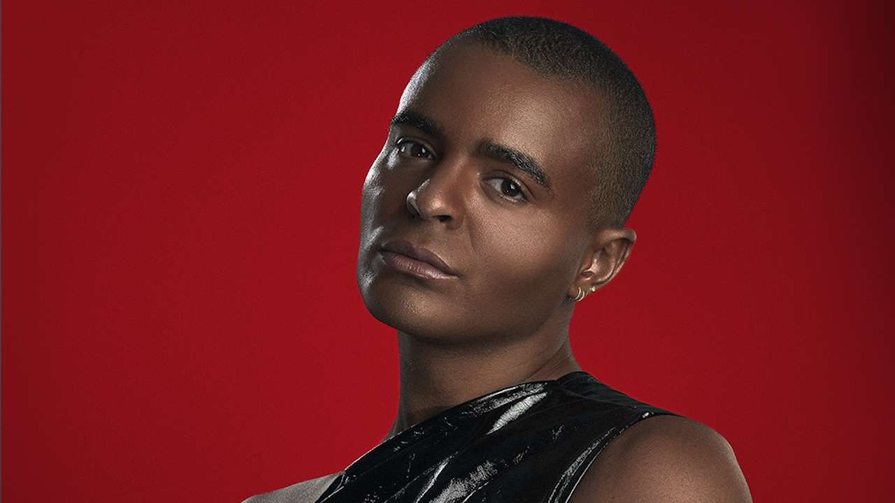 Portrait of a person with a shaved head wearing a black glossy sleeveless top against a red background, neutral expression.