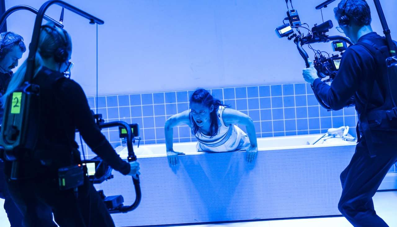 A swimmer climbs over the pool edge as a film crew shoots with large cameras in a blue-lit studio setting.