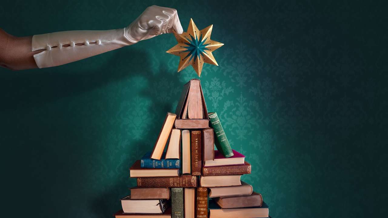 Gloved hand placing a gold star on top of a pyramid of vintage books arranged like a Christmas tree.
