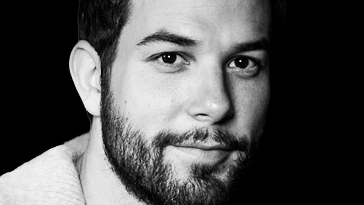 Close-up black-and-white portrait of a man with a beard, looking at the camera with a subtle smile.