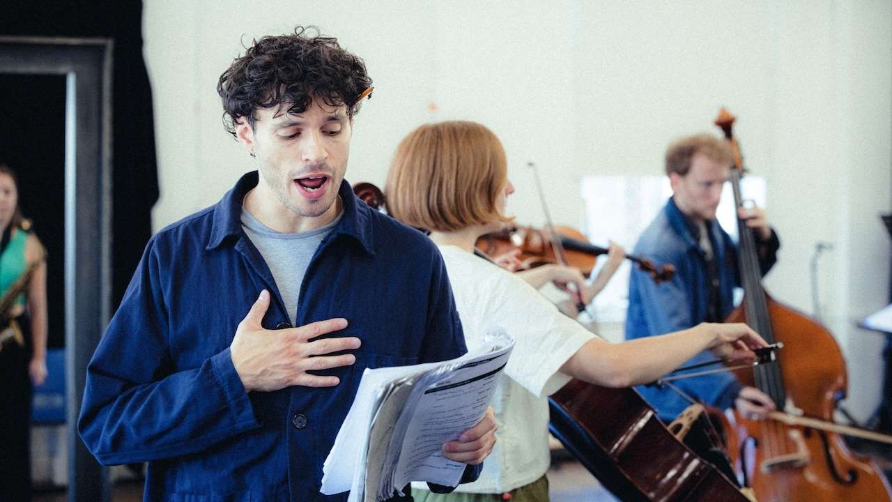 Man in a blue shirt sings with hand on his chest, holding sheet music, as string players rehearse behind him.