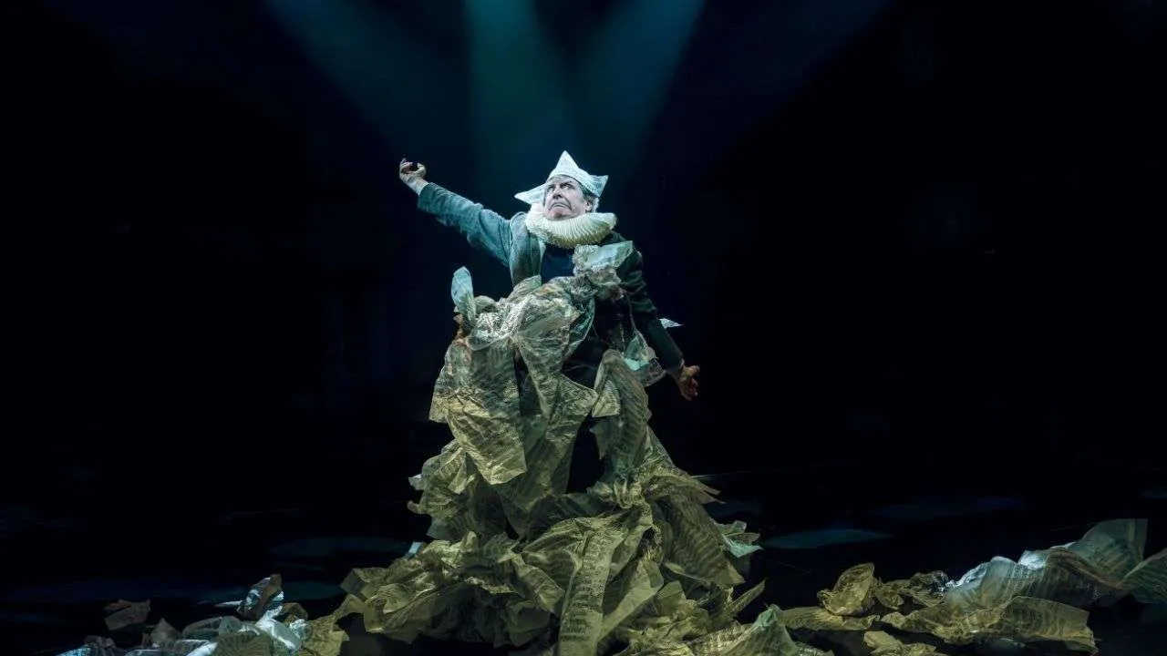 Actor in a crumpled-paper costume on a dark stage, reaching toward the light under a spotlight.