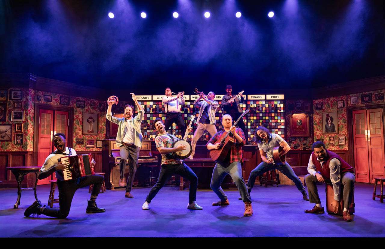 Lively theater stage with a dozen musicians playing guitars, banjo, accordion, and more in a colorful retro set.