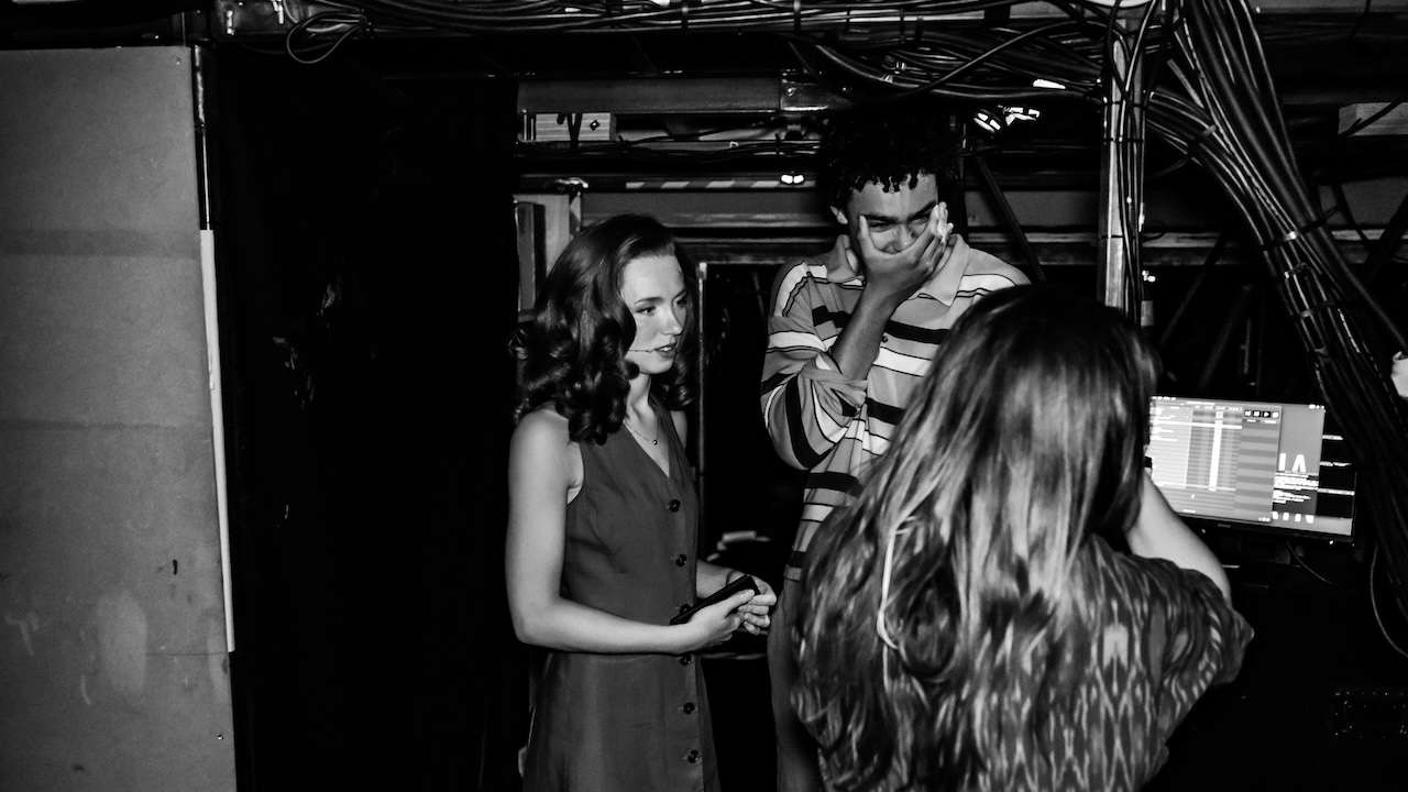 Backstage scene with a woman in a sleeveless dress talking to a man who covers his face, while another person works at a computer console nearby. Cable clutter overhead suggests a tech setup for a performance.