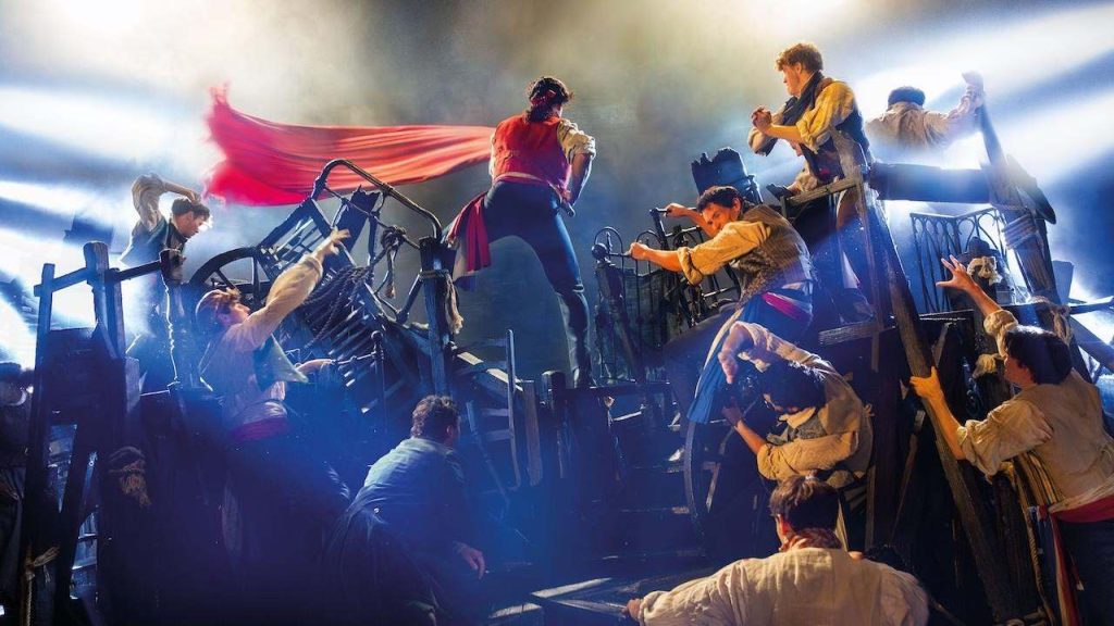 Group of people on a chaotic barricade during a battle scene, dramatic stage lights and a red flag fluttering above them.