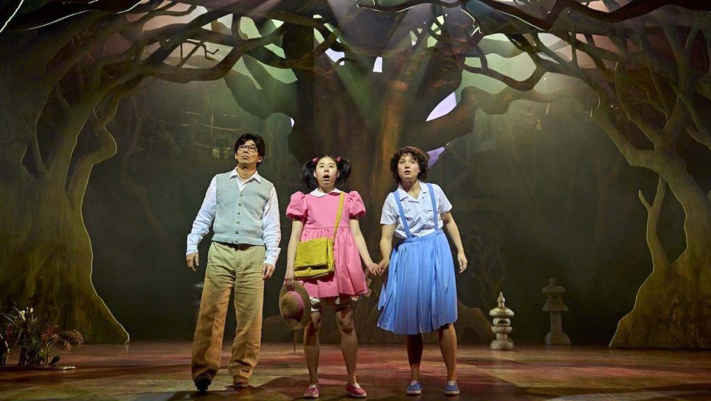 Three children on a theater stage in a forest set, holding hands and looking up.