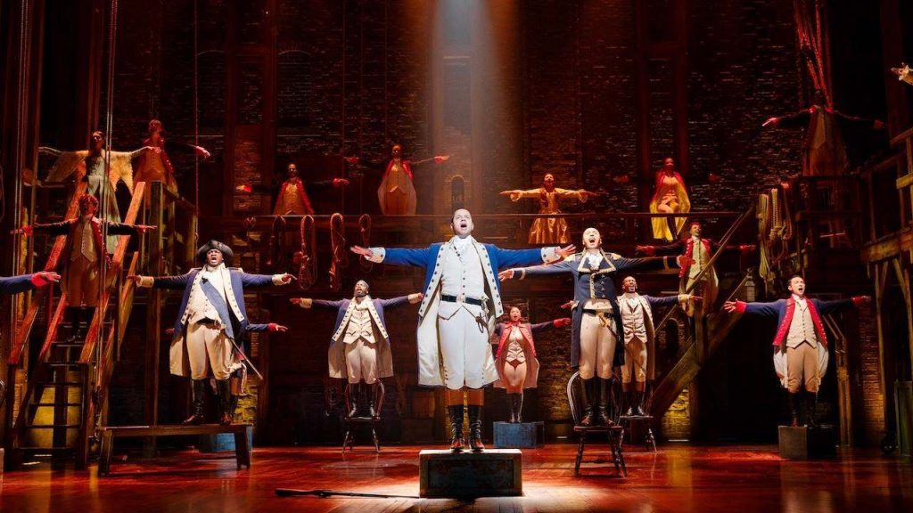 Lead performer on a small raised block with arms wide, surrounded by dancers in 18th‑century costumes on a wooden stage.