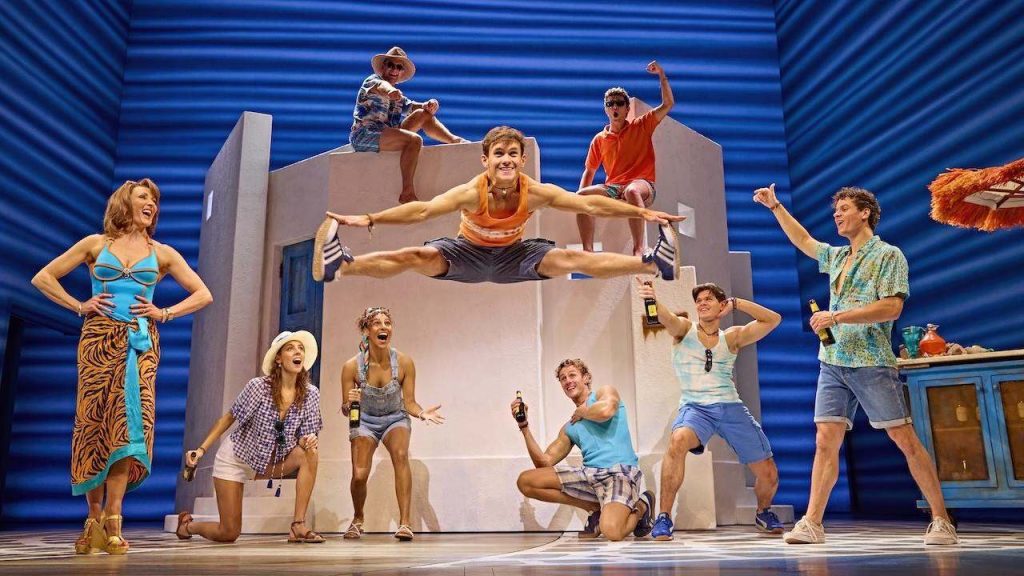 Group of friends on a theater set jump mid-air, celebrating with bottles in hand against a bright blue backdrop.
