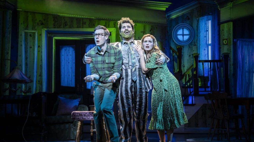 Three actors on a theater stage, a man in a plaid shirt, a man in striped pajamas, and a woman in a green dress, all looking upward with concern.