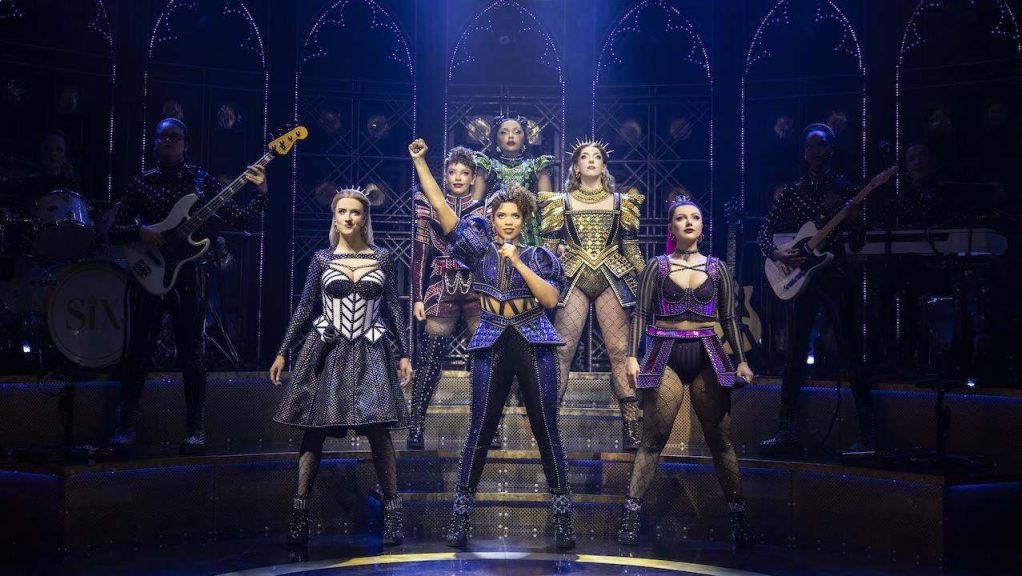 Six performers in metallic, armor-inspired outfits stand on a lit, multi-level stage as a live band plays behind them under blue lighting, mid-performance.