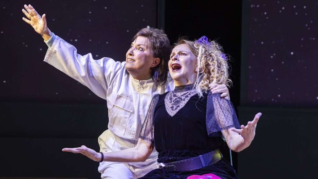 Two performers on stage: a person in a white jumpsuit raising an arm upward while the other in a black dress gestures dramatically, both singing or acting together.