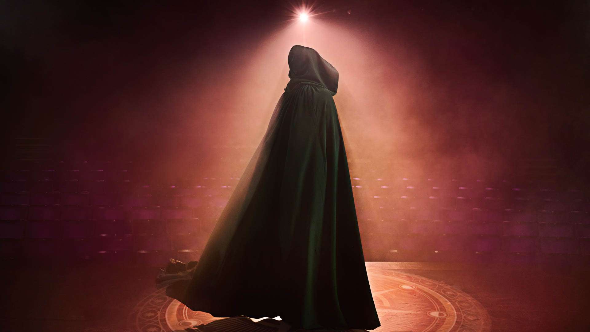 Hooded figure in a dark cloak standing center stage under a bright spotlight, red-tinged background and circular floor pattern visible around them.