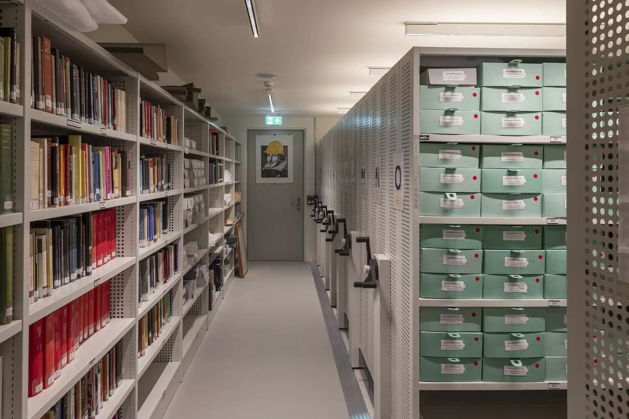 Interior of Shakespeare's Globe's climate-controlled archive store, with rows of bound books on open shelving beside sliding mobile stacks of green archival storage boxes.