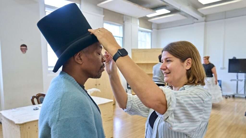 Woman adjusts a tall black top hat on a man's head in a bright studio.