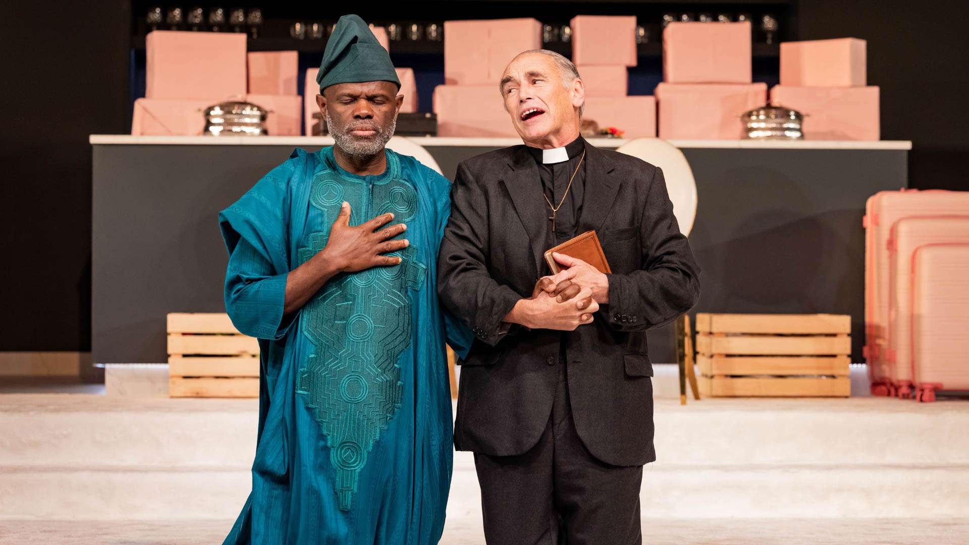 Two men stand close on a stage: one in a teal traditional robe with hand on his chest, the other a priest in a black suit holding a Bible and speaking or singing. Behind them are pink boxes and stage props.