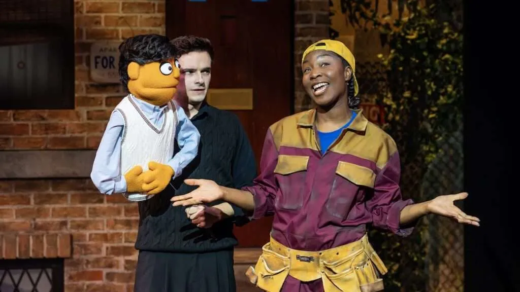 On a brick-stage set, a puppeteer holds an orange puppet wearing a light blue shirt and white vest while a performer in a yellow cap and maroon utility suit gestures with open arms.