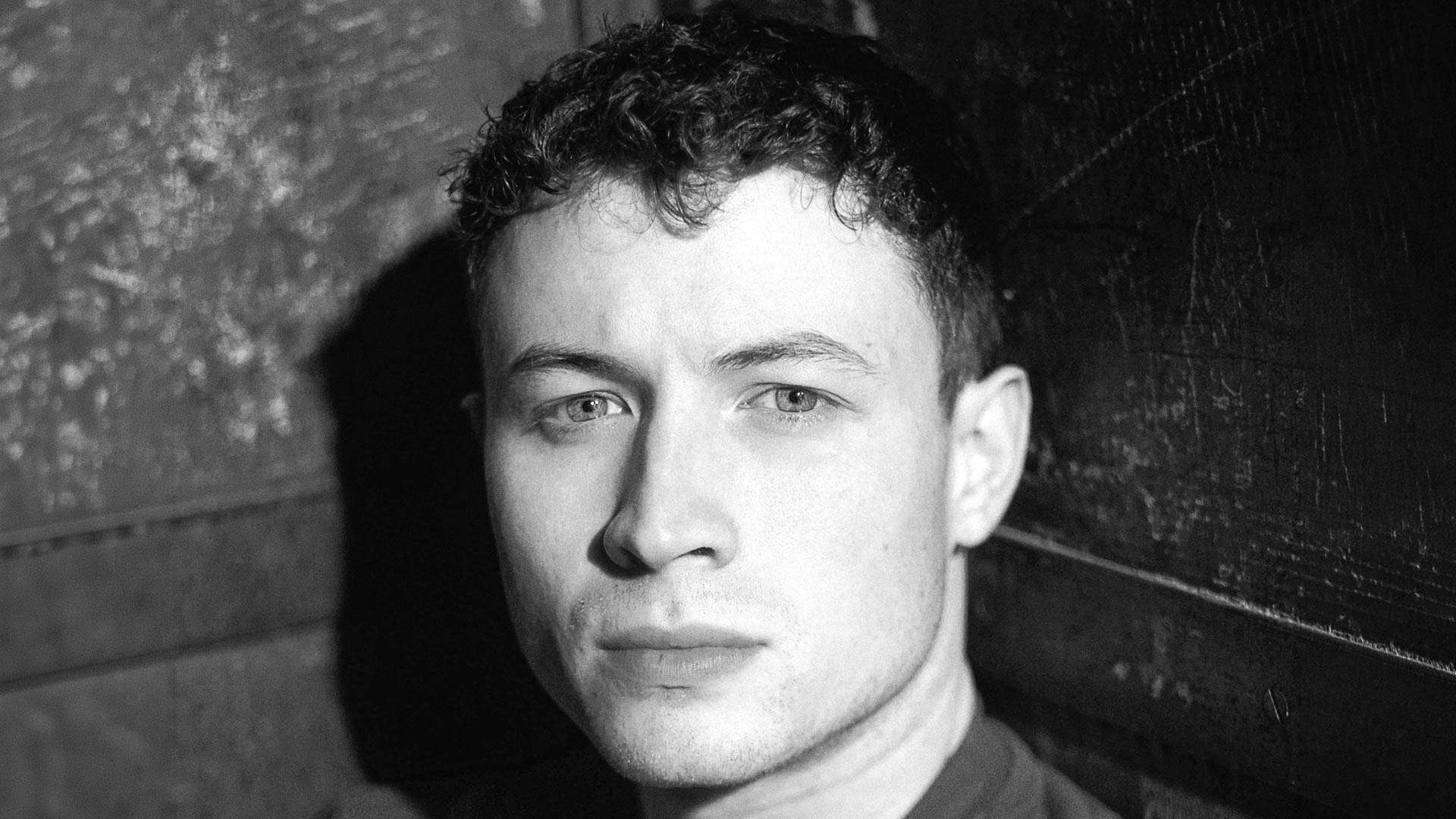 Close-up black-and-white portrait of a young man looking at the camera against a dark wooden background.