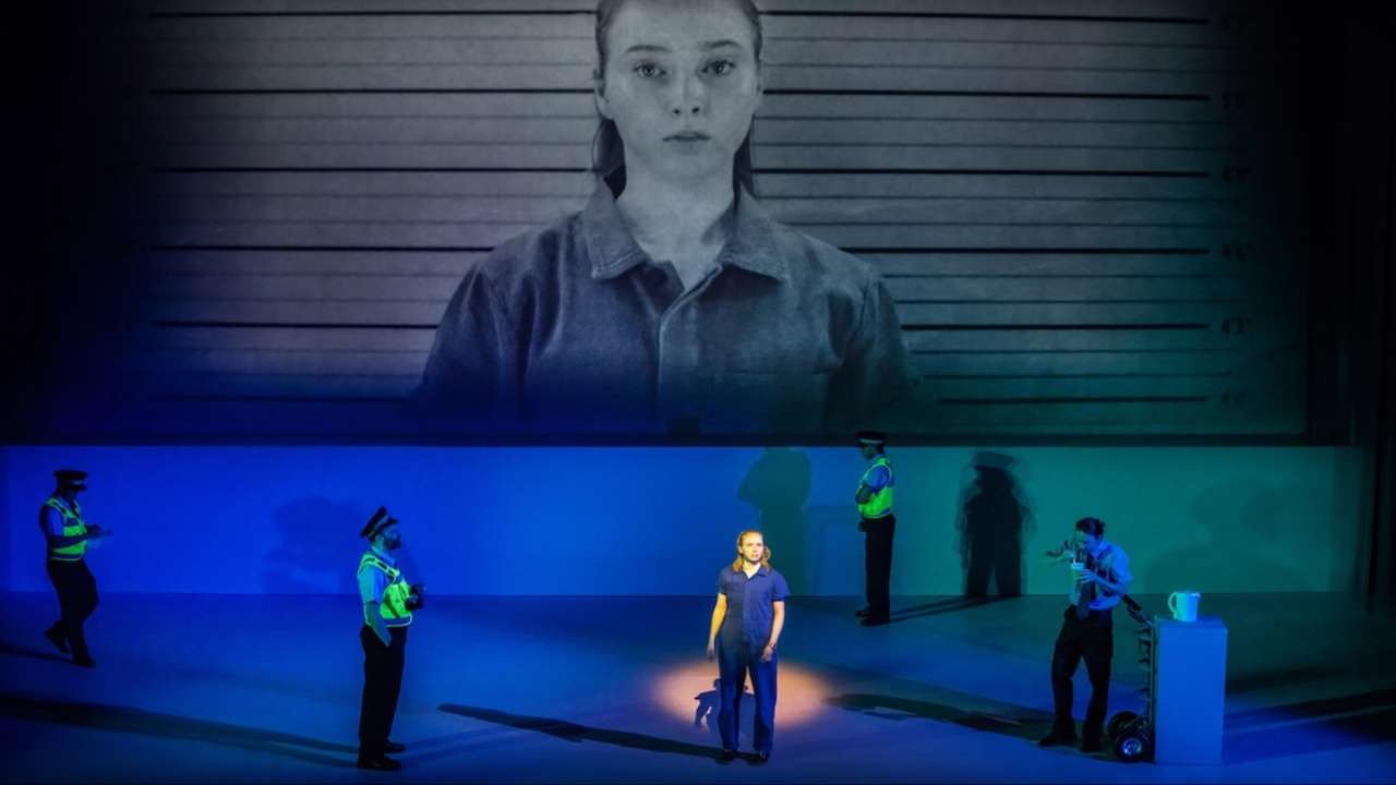 Chris Boone stands alone on stage lit in blue, with a large mugshot-style projection above and police officers positioned around her, in The Curious Incident of the Dog in the Night-Time.