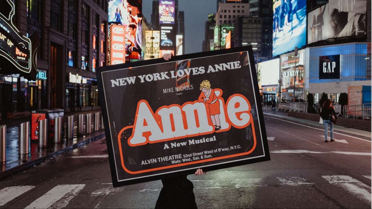 Person walks in a nighttime city street holding a large sign advertising the musical Annie in Times Square, NYC with bright billboards in the background.