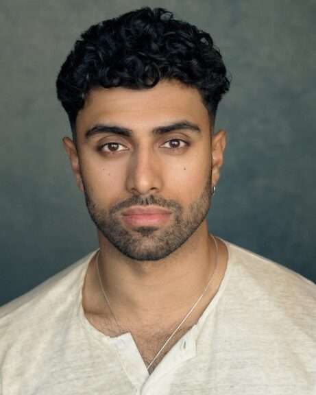 Close-up portrait of a young man with dark curly hair, light stubble, and a small hoop earring, facing the camera.
