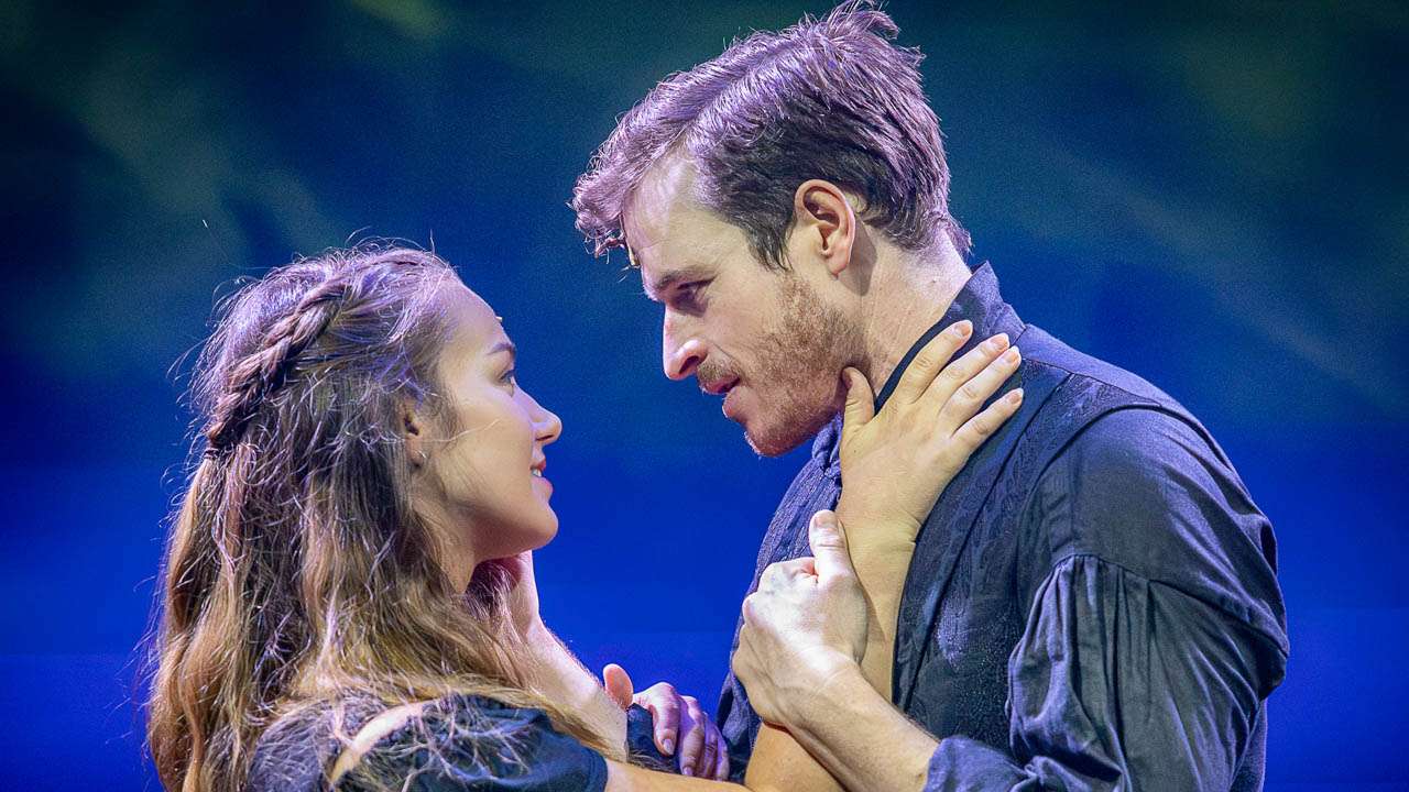 A man and a woman gaze into each other's eyes, holding each other close as if dancing on a stage with blue lighting behind them.