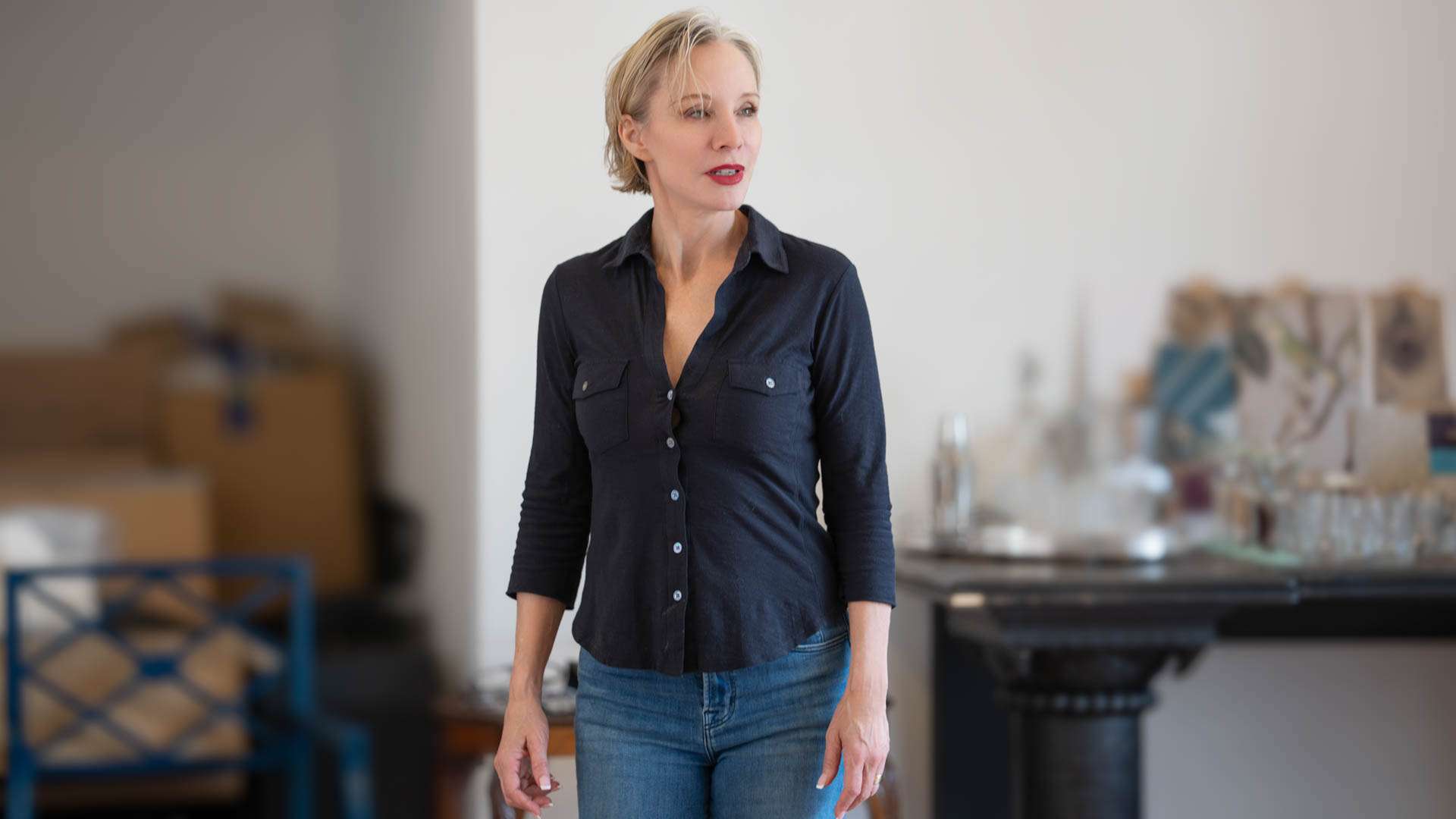 Woman with short blond hair wearing a dark button-down shirt and jeans stands in a cluttered workshop or storage area, looking to the side.