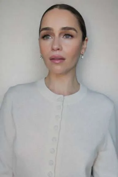 Close-up portrait of a woman with light makeup wearing a white buttoned cardigan and diamond earrings against a neutral background.
