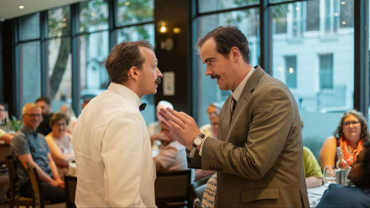 Two men in formal attire converse at a social event, one gesturing with his hands as they face each other.