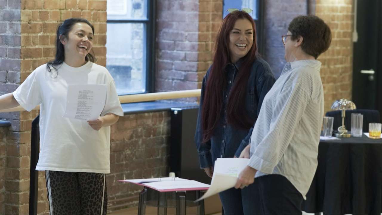 Three women laughing together during a rehearsal, holding script pages, in a brick-walled studio with a cocktail bar setup nearby