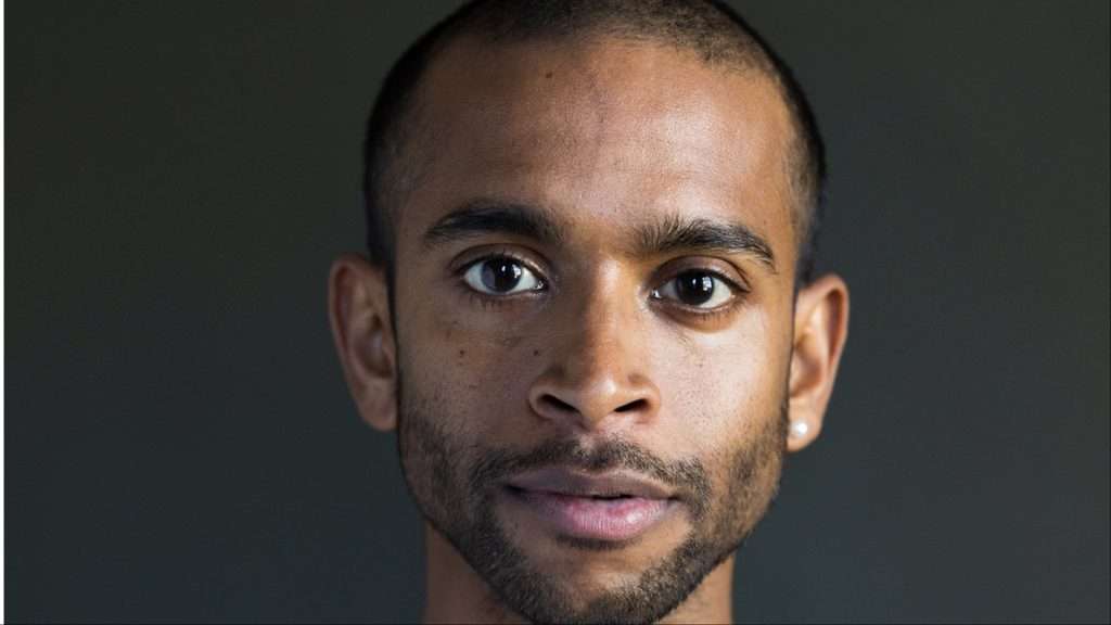 Close-up portrait of a man with short hair and light stubble, looking directly at the camera against a dark background.