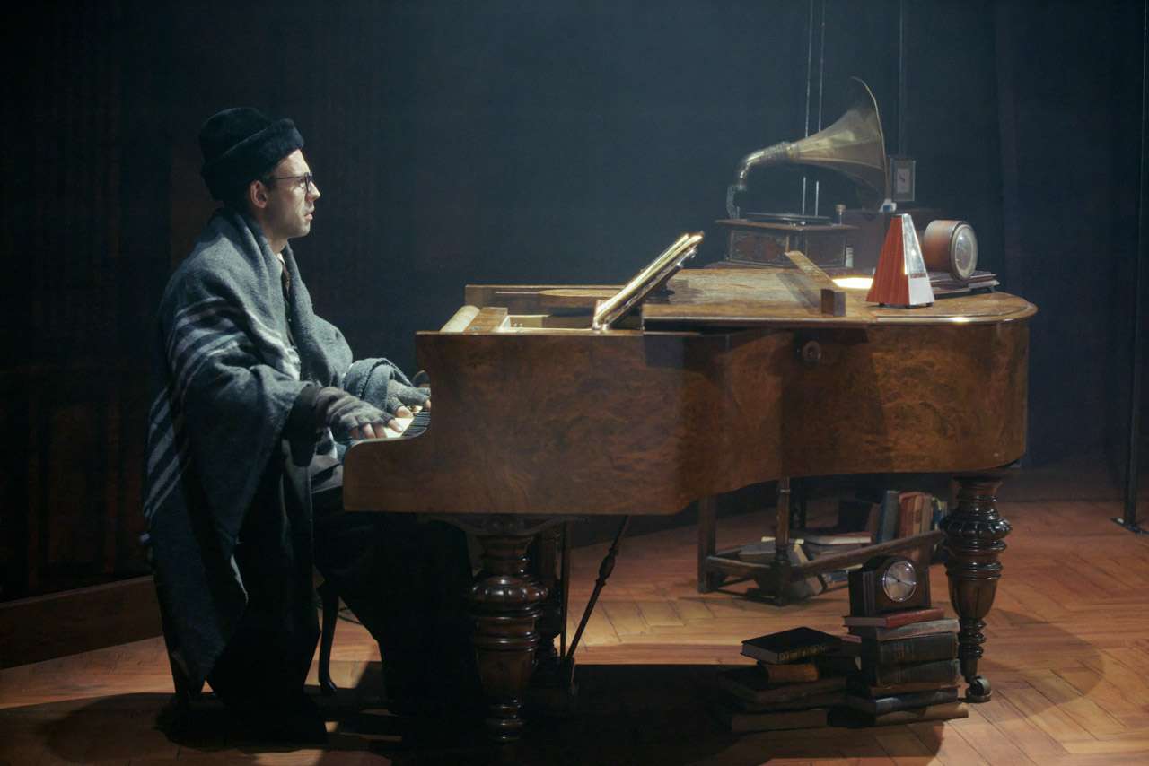 Pianist in vintage clothing sits at an old wooden piano on a dim stage, with stacked books nearby and a gramophone on the piano lid.