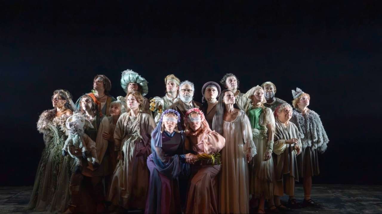 The company of Into the Woods stand together in muted fairytale costume against a black backdrop, two brides at the front with floral blindfolds.