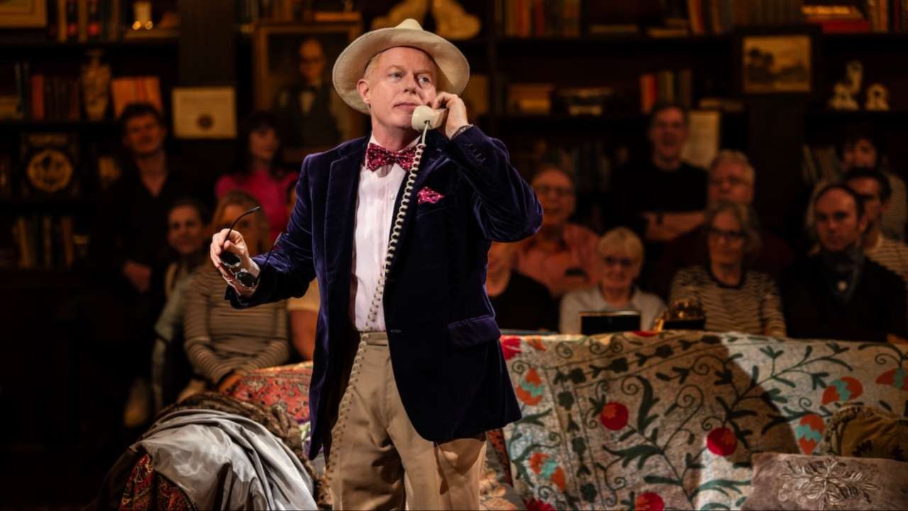 Jesse Tyler Ferguson as Truman Capote in Tru, wearing a navy velvet jacket, straw hat and bow tie while speaking on a corded telephone, with the audience seated behind him on stage.