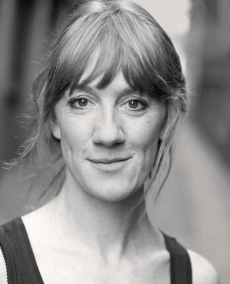 Close-up black-and-white portrait of a smiling woman with light hair and subtle freckles, wearing a dark tank top.