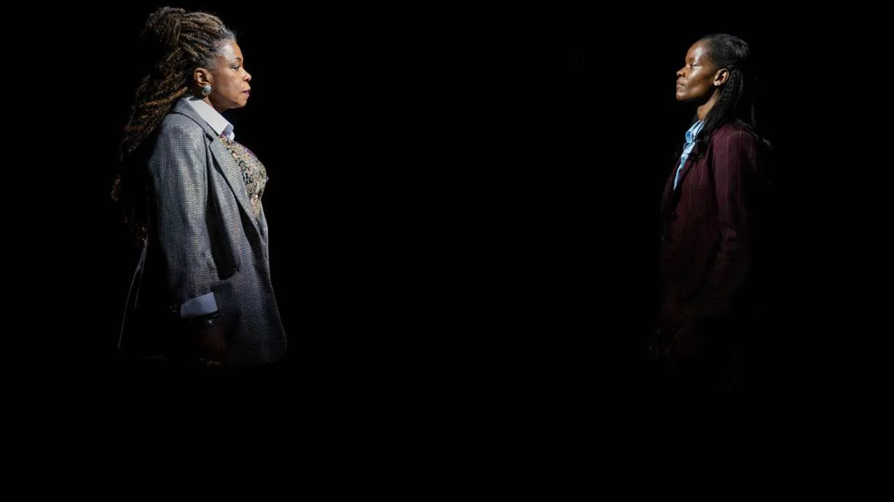 Two women in business attire stand apart, facing each other in a dark, spotlighted setting.