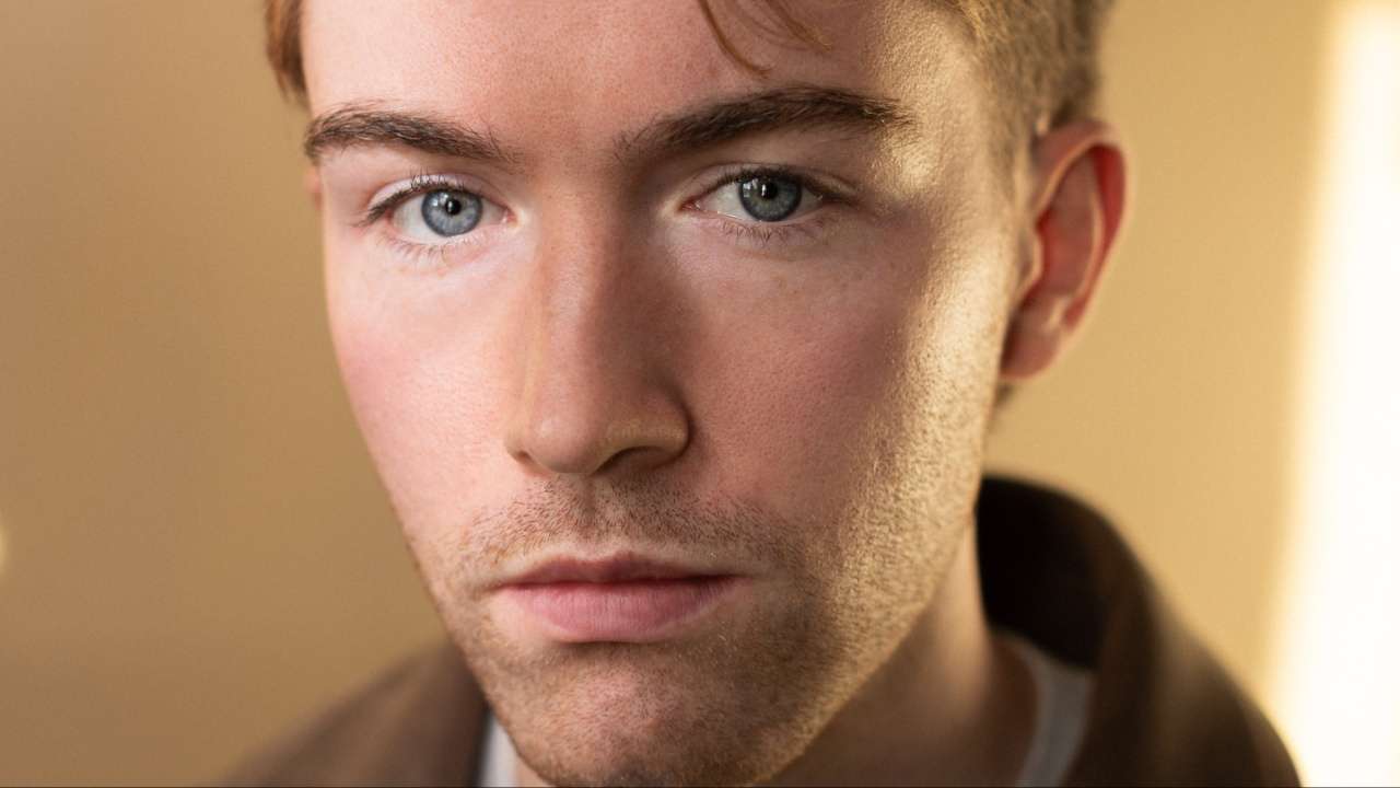 Close-up portrait of a young man with blue eyes looking at the camera, neutral expression, warm lighting.