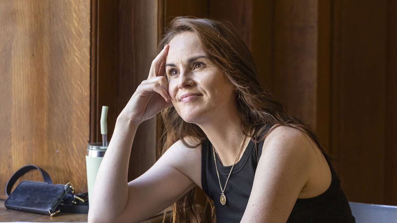 Woman sits at a wooden table, resting her head on her hand with a gentle smile, indoor light nearby.