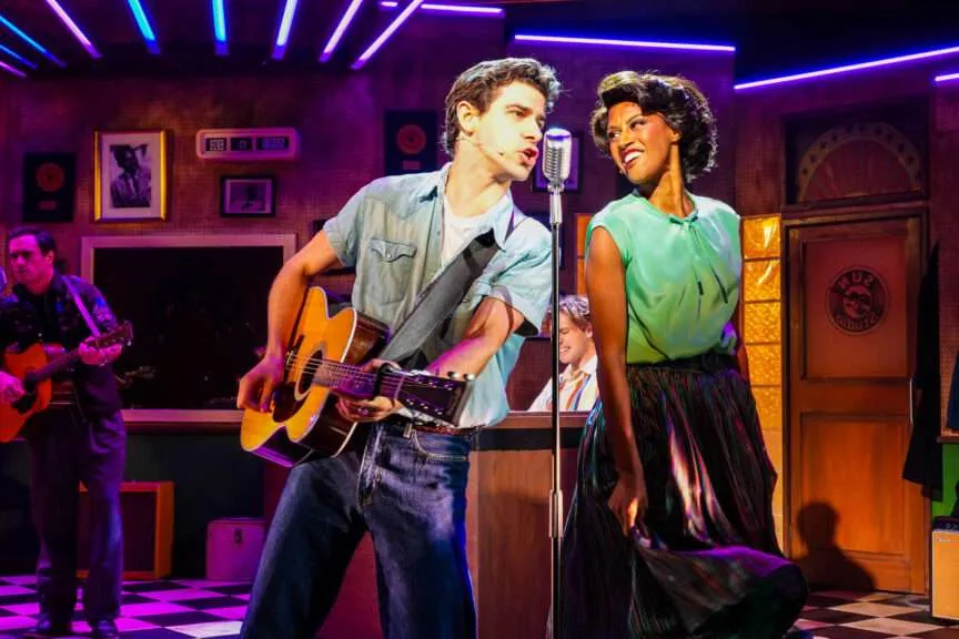 Musician duo on stage: a man with a guitar singing into a microphone while a woman in a green blouse harmonizes beside him, with neon purple lights and a retro lounge backdrop behind them.