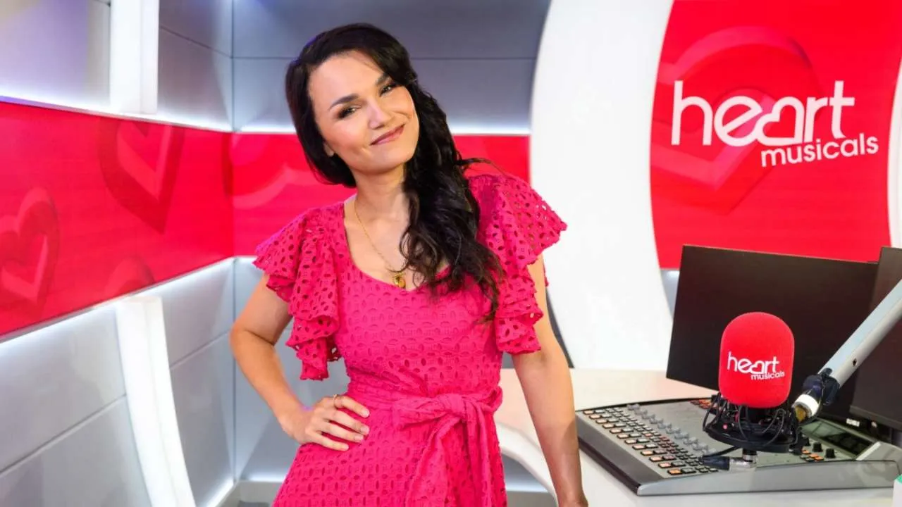 Samantha Barks in a pink dress posing at a Heart Musicals studio desk with a red microphone haloed by the logo behind her.