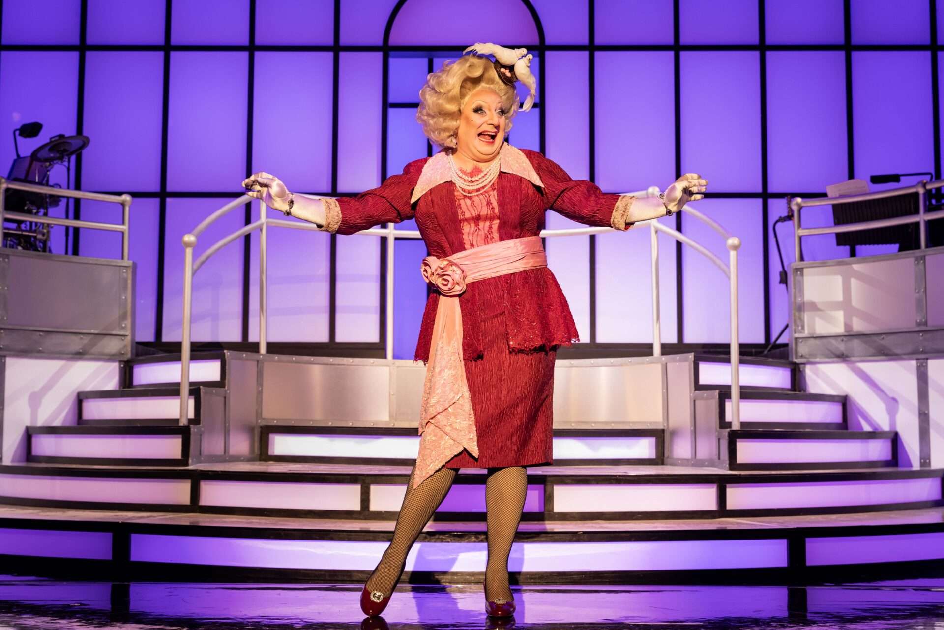 Drag queen performer in a red embellished dress with a pink sash, posing with arms outstretched on a lit theater stage with purple backlighting.