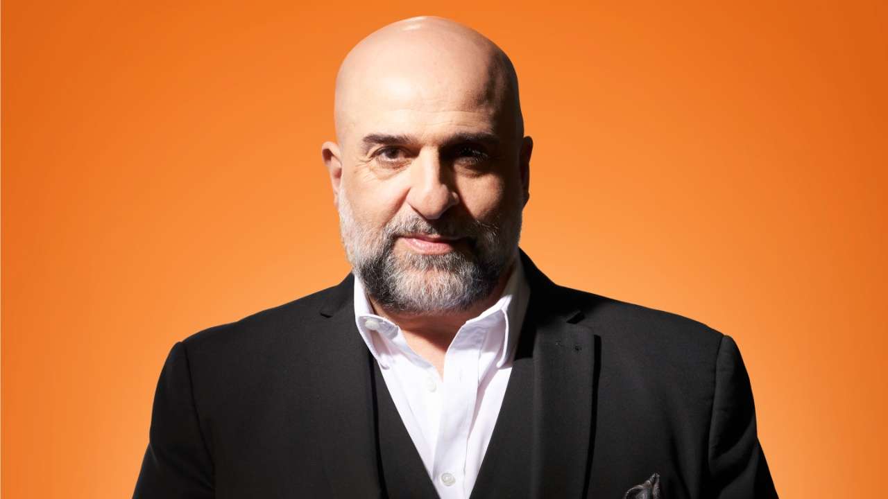 Omid Djalili in a suit in front of orange background