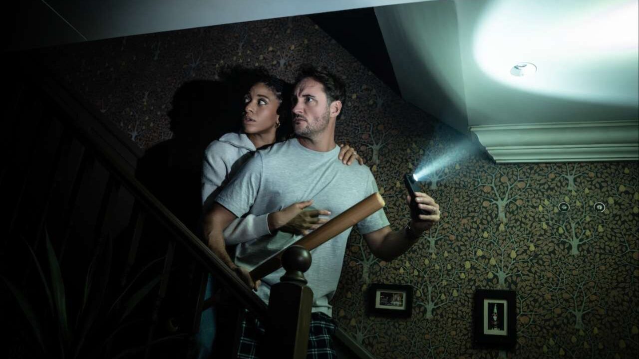 Production photo from Paranormal Activity showing two cast members on a staircase using a phone torch amid the dark.