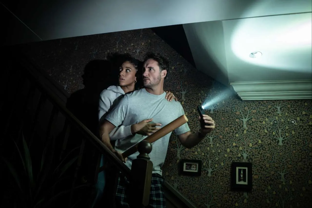 Production photo from Paranormal Activity showing two cast members on a staircase using a phone torch amid the dark.